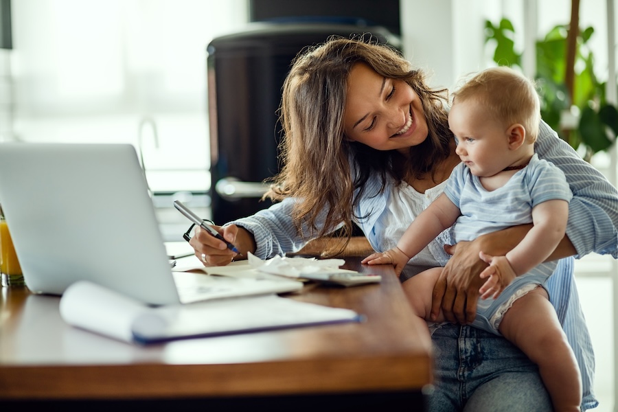 A parent holding a baby on one hip while working through the household budget at a laptop