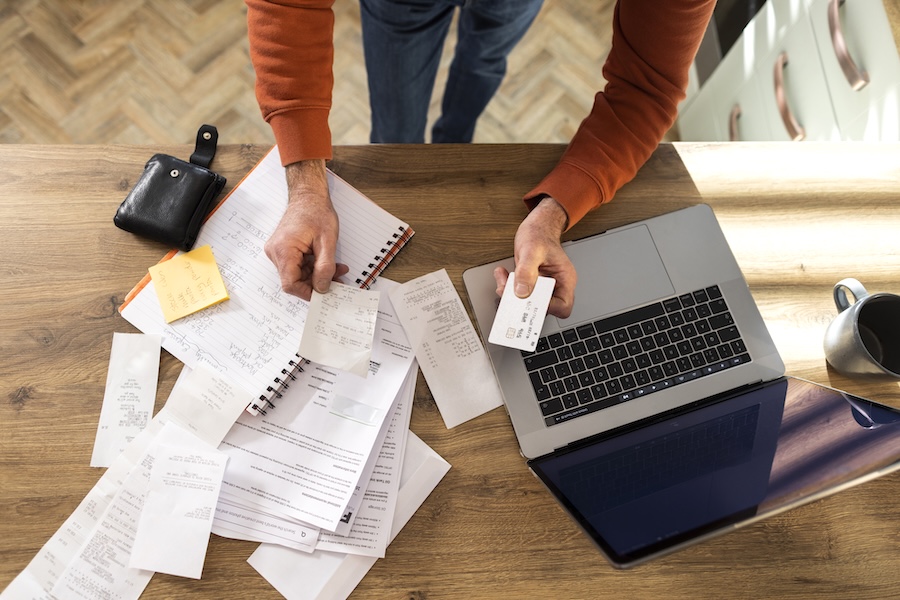 A table covered in receipts and handwritten notes beside a laptop and a debit card