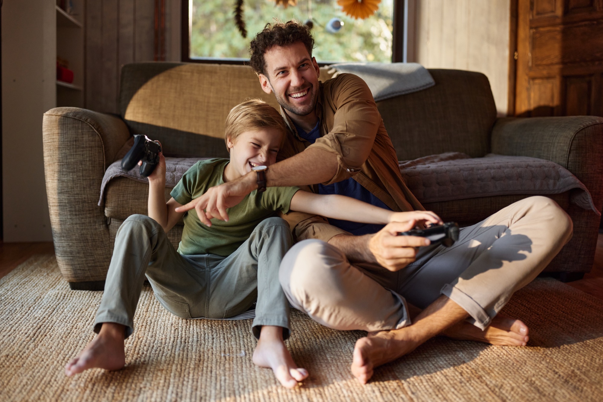 A father and son laughing while playing video games together on the living room floor