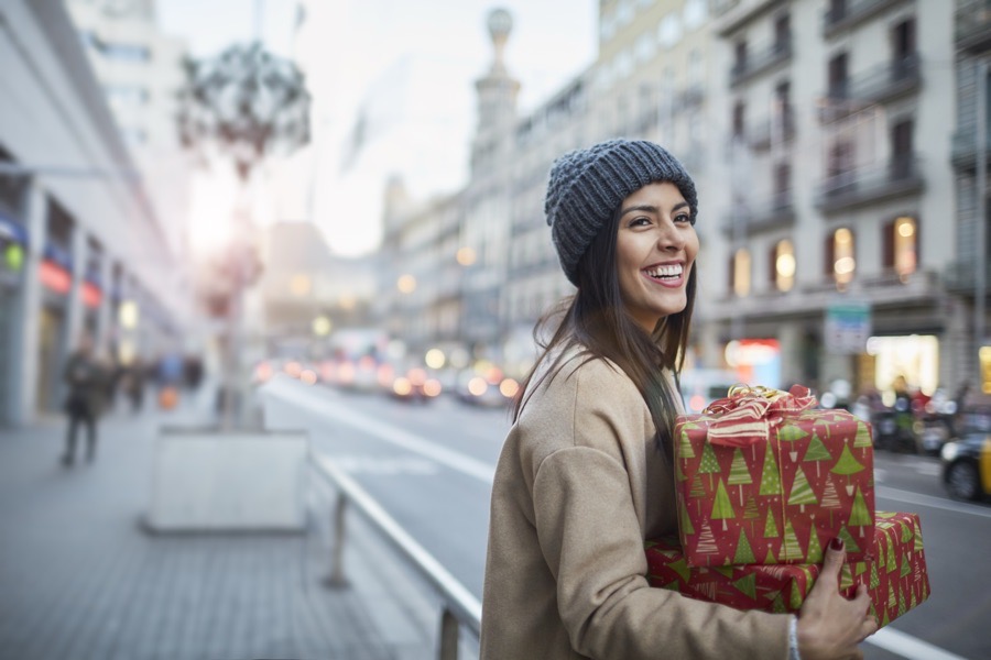 A smiling woman on a city street carrying a stack of wrapped holiday gifts