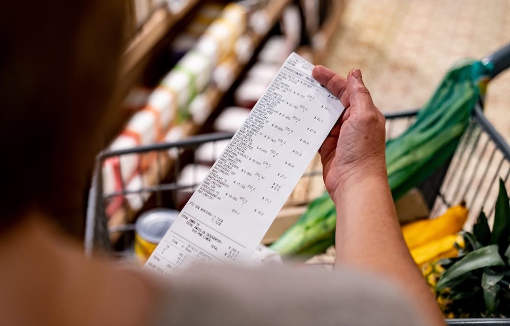 A hand holding a long grocery receipt over a shopping cart full of groceries