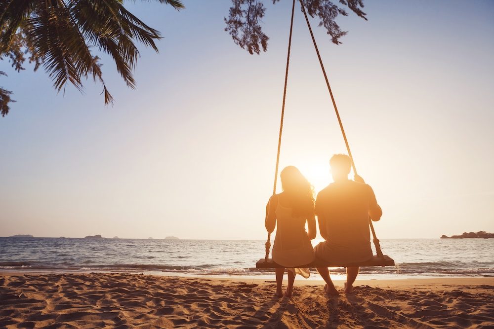 A couple sitting together on a swing on the beach at sunset